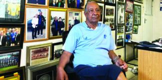 Augusto Palacios sitting in his home study surrounded by football trophies and memorabilia